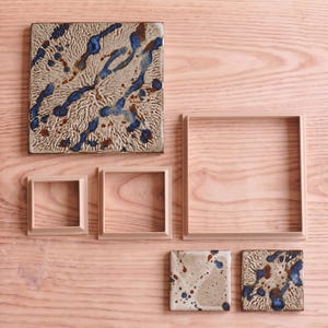 May include: A collection of square ceramic tiles and matching square frames. One large tile features a textured beige surface with blue and brown splatters. Two smaller tiles have similar speckled patterns. Three empty square frames of varying sizes are included.