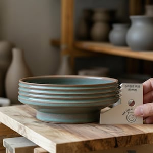 Op de afbeelding: Een handgemaakte, turquoise keramische kom met horizontale ribbels. De kom staat op een houten oppervlak, en een gereedschap met de tekst "GP007 RIB 80mm" wordt ernaast gehouden. De kom heeft een bruine rand en bodem.