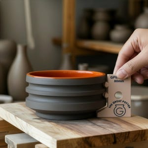 Peut inclure: Un bol en céramique grise avec un intérieur orange. Le bol a un extérieur strié et est mesuré avec un outil indiquant 80 mm. Le texte "THE GREAT POTTER" et un logo sont visibles sur l'outil. Le bol est posé sur une surface en bois.