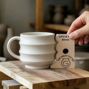 Pode incluir: Uma caneca de cerâmica branca com um design canelado, ao lado de uma ferramenta bege com o texto "THE GREAT POTTER" e "GP018 R 80mm". A caneca está sobre uma superfície de madeira, com objetos de cerâmica ao fundo.