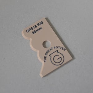 Puede incluir: Una herramienta de alfarer&iacute;a beige con borde festoneado, un orificio central circular y texto impreso en negro que dice GP018 RIB 80mm y THE GREAT POTTER con un logotipo circular. La herramienta tiene una forma plana y angular para modelar arcilla.