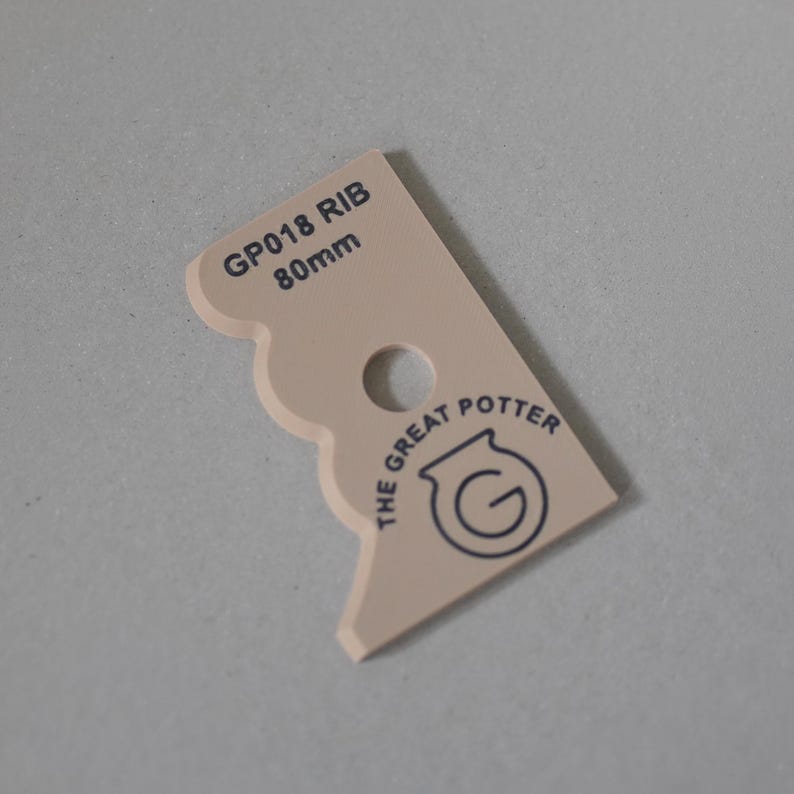Puede incluir: Una herramienta de alfarer&iacute;a beige con borde festoneado, un orificio central circular y texto impreso en negro que dice GP018 RIB 80mm y THE GREAT POTTER con un logotipo circular. La herramienta tiene una forma plana y angular para modelar arcilla.