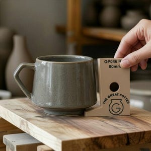 May include: A gray ceramic mug with a geometric handle sits on a wooden surface. A beige measuring tool with the text "GP046 R 80mm" and "THE GREAT POTTER" is held nearby. The mug has a wide base and a slightly textured surface.