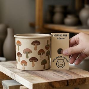 Puede incluir: Una maceta de cerámica beige con un patrón repetido de setas coloridas. La maceta es cilíndrica. Una tarjeta junto a la maceta dice "GP003 RIB 80mm". La maceta mide unos 8 cm de alto.