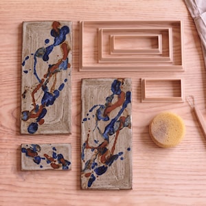 May include: Two rectangular ceramic tiles with a speckled beige base and blue and brown paint splatters, three nested rectangular plastic frames, one small rectangular plastic frame, a round yellow sponge, and a metal wire clay tool.