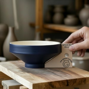 Può includere: Una ciotola in ceramica blu navy con interno color crema è appoggiata su una superficie di legno. La ciotola ha una forma svasata e viene misurata con uno strumento che indica "GP026 RIP 80mm". Il logo The Great Potter è visibile.