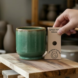 Puede incluir: Una taza de cerámica verde con textura acanalada y borde marrón. La taza está junto a una herramienta de medición beige que dice "GP023 RIP 100mm" y "THE GREAT POTTER". La taza está sobre una superficie de madera.