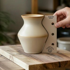 May include: Cream ceramic vase with a flared rim and a curved body. The vase has a brown band around the middle and top. A white measuring tool with the text "GP028 RIB 200mm" is next to the vase. The vase is on a wooden surface.