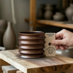 Può includere: Vaso in ceramica marrone con design a coste. Il vaso è cilindrico e poggia su una superficie di legno. Uno strumento beige con la scritta "GP032 RIB 100mm" è tenuto accanto al vaso. È visibile anche il logo The Great Potter. Altezza di circa 10 cm.