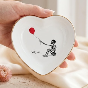 Op de afbeelding: Een wit hartvormig keramieken schaaltje met een goudkleurige rand. Het midden bevat een zwarte illustratie van een zittend skelet dat een rode ballon vasthoudt, vergezeld van de zwarte gedrukte tekst die luidt, Well, shit...