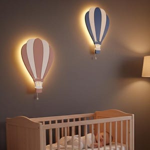 May include: A child's bedroom with a light wood cot and a sleeping baby. Two hot air balloon wall lights, one pink and one blue, are mounted on a dark grey wall. A grey armchair with a blanket and toy is also visible.