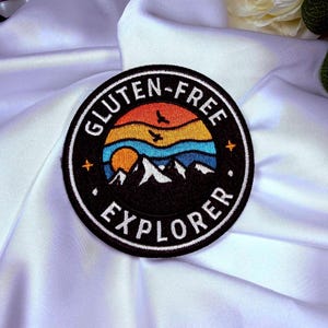 Peut inclure: Un écusson rond noir avec l'inscription blanche "GLUTEN-FREE EXPLORER". Le patch présente un motif coloré de montagnes, de coucher de soleil et d'oiseaux. Il s'agit probablement d'un écusson à repasser ou à coudre.