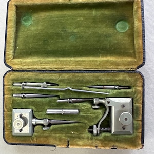 May include: A vintage metal drafting instrument set housed in a rectangular case with a dark exterior and a green velvet interior. The set includes various steel components, including pointed tips, a central rod, and two mechanical adjustment blocks.