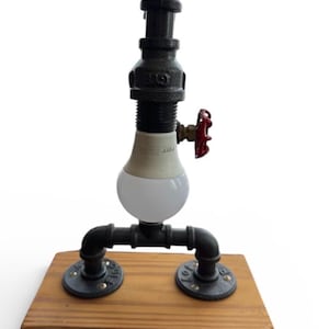 Puede incluir: Lámpara de mesa de estilo industrial hecha de tuberías y accesorios negros, con una bombilla redonda y blanca. La lámpara tiene un mango de válvula rojo y está montada sobre una base de madera rectangular. La altura de la lámpara es de unos 30 cm.