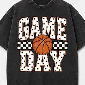 May include: A charcoal black t-shirt featuring a graphic print with the text GAME DAY in large white block letters with black polka dots. A central orange basketball is positioned between the words, set against a black and white checkered stripe.