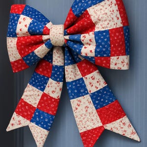 May include: A large, decorative bow in a patchwork design. The bow features a pattern of red, white, and blue squares, with a floral print on the white fabric. The bow is likely made of fabric and is suitable for patriotic or holiday decor.