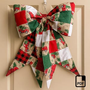 May include: A large, decorative bow made of quilted fabric, featuring a patchwork design with red trucks carrying Christmas trees, red and black plaid, green squares, and white accents. The bow is suspended from a metal hook, perfect for holiday decor.