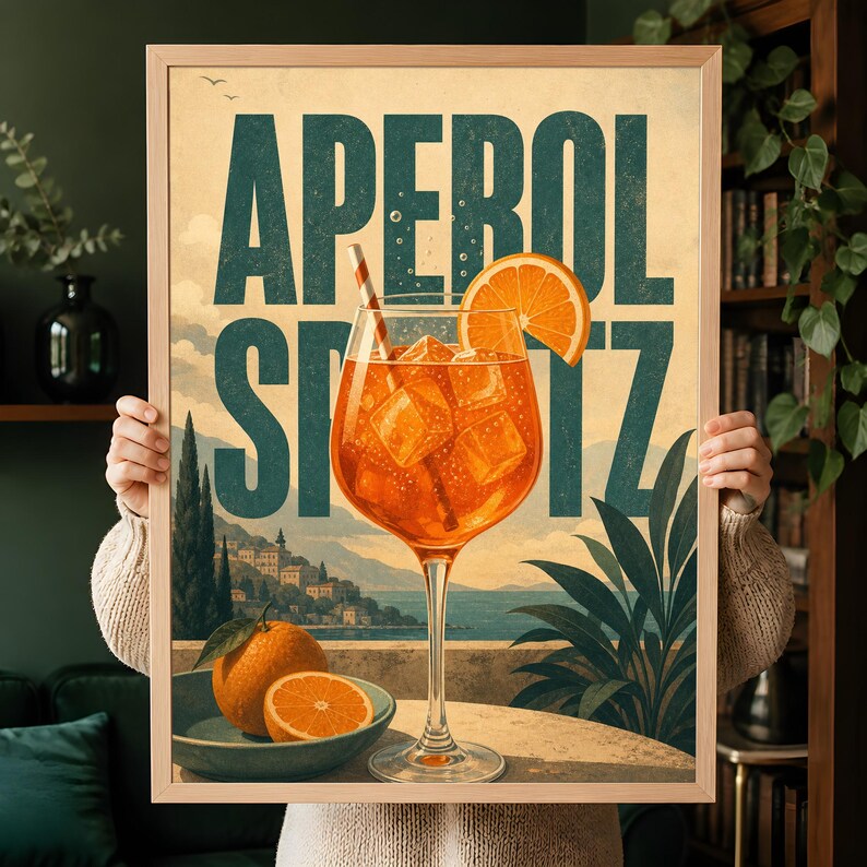 Puede incluir: Un p&oacute;ster enmarcado con el texto APEROL SPRITZ en letras grandes de color verde azulado oscuro. La ilustraci&oacute;n muestra una copa con un c&oacute;ctel naranja, cubitos de hielo y una rodaja de naranja, frente a un paisaje costero y un cuenco de naranjas.
