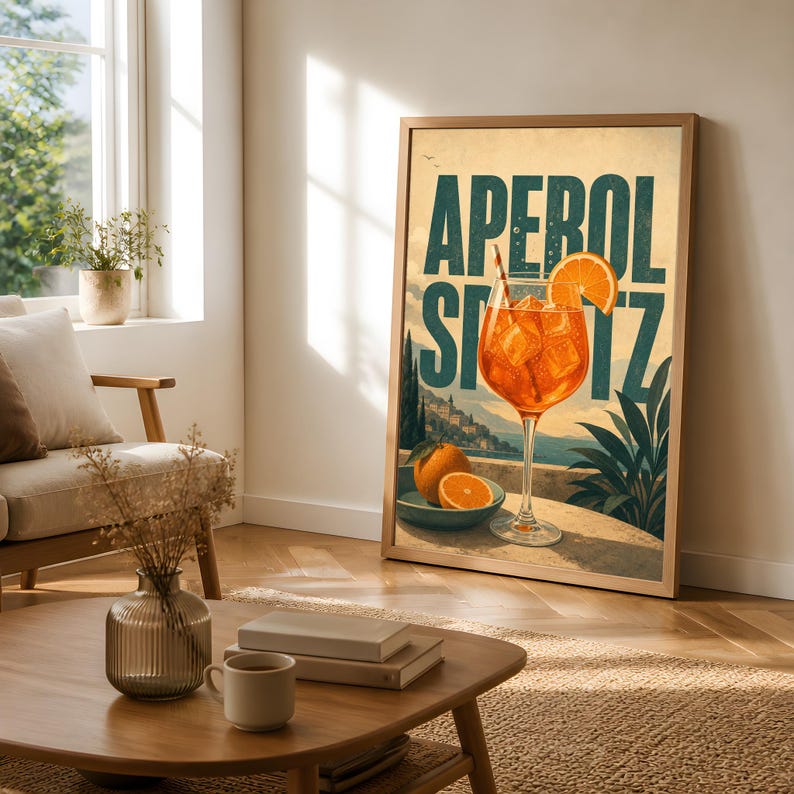 Puede incluir: Un cuadro vertical enmarcado que presenta una ilustraci&oacute;n de estilo vintage de un c&oacute;ctel Aperol Spritz en una copa con hielo y una rodaja de naranja, sobre un paisaje costero con el texto en negrita APEROL SPRITZ.