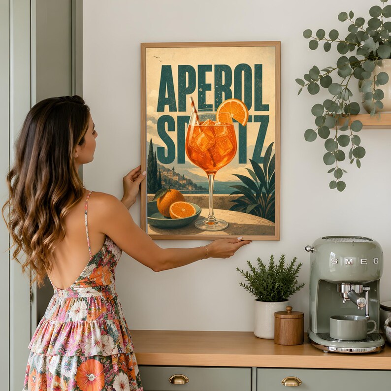 Puede incluir: Un p&oacute;ster de pared rectangular que presenta una ilustraci&oacute;n vibrante de un c&oacute;ctel Aperol Spritz en una copa de tallo, con rodajas de naranja y un paisaje costero. El texto APEROL SPRITZ est&aacute; impreso en letras may&uacute;sculas grandes, negritas y azul verdoso.