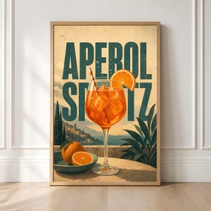 Puede incluir: Un p&oacute;ster enmarcado que muestra un c&oacute;ctel Aperol Spritz de color naranja vibrante en una copa con hielo y una pajita. El texto APEROL SPRITZ aparece en letras grandes de color verde oscuro. Un cuenco con naranjas enteras y cortadas descansa sobre una mesa.