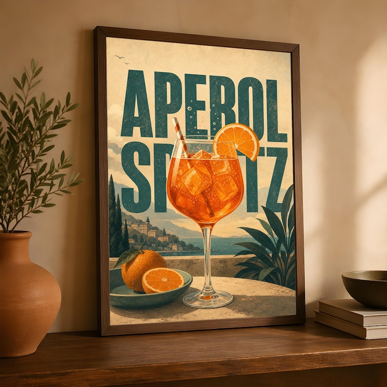 Puede incluir: Una l&aacute;mina enmarcada que muestra un vaso alto de Aperol Spritz naranja con cubitos de hielo y una pajita, sobre una mesa con naranjas frescas. El fondo muestra un paisaje costero con el texto APEROL SPRITZ en letras de color azul verdoso.