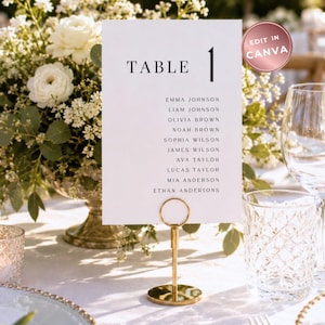 May include: A white rectangular wedding table seating chart card with black text reading TABLE 1 followed by a list of names including Emma Johnson, Liam Johnson, Olivia Brown, Noah Brown, Sophia Wilson, James Wilson, Ava Taylor, Lucas Taylor, Mia Anderson, and Ethan Anderson, held by a gold circular stand.