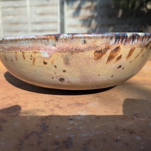 May include: A shallow ceramic bowl with a rounded profile, featuring a variegated glaze in shades of tan, beige, and brown. The rim displays a darker, mottled brown and blue-toned finish with visible drips and speckles across the exterior surface.