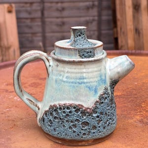 May include: A ceramic teapot featuring a light blue and white glazed upper section and a dark, textured, cratered blue-grey base. The teapot includes a matching lid with a central knob, a curved handle, and a short, angled spout.