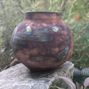May include: A round, wide-mouthed ceramic vase with a textured, matte surface. The exterior features a mottled blend of deep brown, copper, and dark charcoal tones, accented by iridescent patches of metallic blue, green, and gold throughout the body.