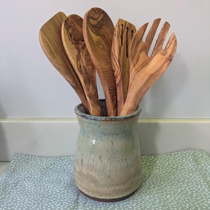 May include: A set of five wooden kitchen utensils with distinct grain patterns, including spoons, a spatula, and a fork, are arranged inside a ceramic crock. The crock features a mottled light blue and beige glaze with a dark rim.