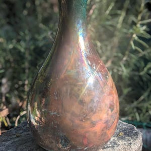 May include: A pear-shaped, iridescent glass vase with a dark green rim. The vase has a metallic sheen with colours of bronze, copper, and hints of blue and purple. The vase is sitting on a grey stone.