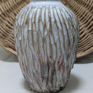 Könnte beinhalten: Eine Keramikvase mit einer texturierten, vertikal gerippten Oberfläche, die mit einer matten weißen Glasur versehen ist. Die tiefen Rillen enthüllen erdige braune und metallisch goldene Untertöne, was für einen starken Kontrast sorgt.