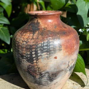 May include: A handmade ceramic vase with a textured finish. The vase displays a mix of earthy colours, such as reddish-brown, black, and cream. The design incorporates a pattern resembling scales or a network. The vase has a rounded body and a slightly flared rim.