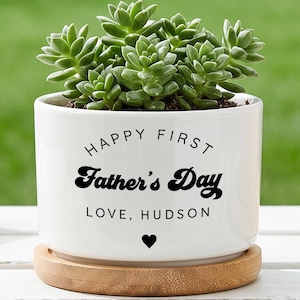 May include: A white ceramic planter with a green succulent plant. The planter has the text "HAPPY FIRST Father's Day LOVE, HUDSON" in black. It sits on a round wooden coaster. A heart symbol is below the text.