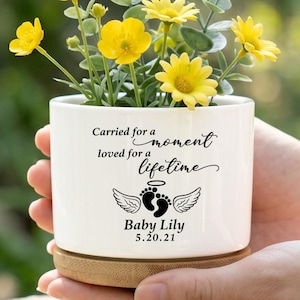May include: A white ceramic planter with a bamboo base containing yellow flowers. The planter features black text reading Carried for a moment loved for a lifetime, an illustration of baby footprints with angel wings, and the name Baby Lily 5.20.21.