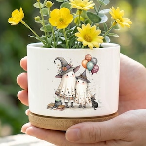 May include: A white ceramic planter with a bamboo saucer containing yellow flowers and green foliage. The planter features a graphic illustration of two ghost figures wearing witch hats, holding balloons, standing near a black cat and books.