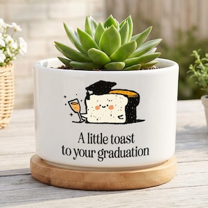May include: A white ceramic cylindrical plant pot sitting on a round bamboo saucer. The pot features a graphic of a piece of toast wearing a graduation cap and holding a glass, with the text A little toast to your graduation below it.
