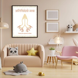 May include: A framed art print with a black border, featuring a gold illustration of hands in prayer and text in a foreign language. The text below reads "Treat your guest as God." The print hangs above a peach-colored sofa.