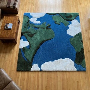 Puede incluir: Una alfombra cuadrada con un diseño de mapa del mundo. Presenta océanos azules, continentes verdes y formas blancas en forma de nube. La alfombra está sobre un suelo de madera.