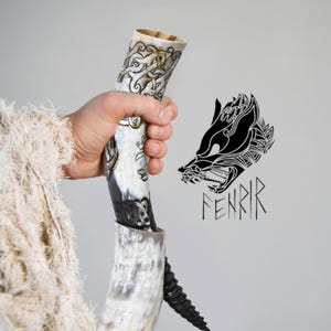 Pode incluir: Um chifre para beber em estilo Viking com intrincados desenhos esculpidos. O chifre é segurado em uma mão e apresenta uma base escura em espiral. Um gráfico de cabeça de lobo em preto e branco com a palavra "FENRIR" está à direita.