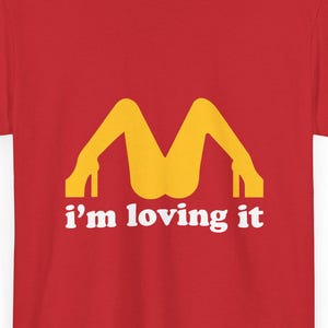 Può includere: T-shirt rosso con grafica gialla di gambe con tacchi alti che formano una "M", con la scritta bianca "i'm loving it" sotto. La maglietta è di colore rosso pieno.
