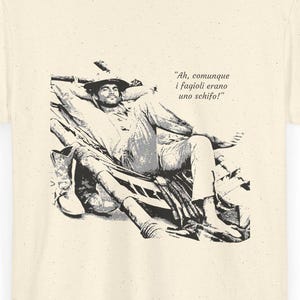 Può includere: T-shirt color crema con un'illustrazione in bianco e nero di un uomo reclinato, con stivali e cappello. Il testo sulla maglietta recita: "Ah, comunque i fagioli erano uno schifo!"