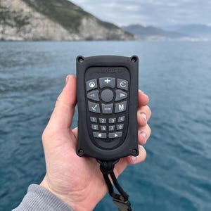 May include: A black MotorGuide remote control with multiple buttons and a lanyard. The remote has a series of numbered buttons, directional arrows, and function buttons. The background shows a body of water and a mountain.