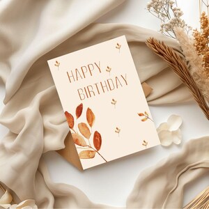 Puede incluir: Una tarjeta de cumpleaños color crema con las palabras "HAPPY BIRTHDAY" en letras marrones. La tarjeta presenta hojas de acuarela en tonos naranja y marrón. La tarjeta está colocada sobre una tela beige con flores secas.