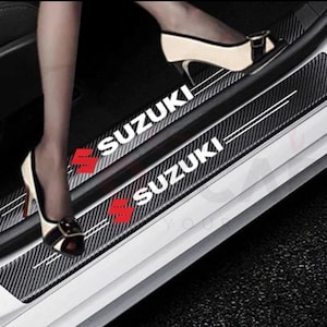 Puede incluir: Protectores de umbral de puerta de coche negros y plateados con el logotipo de Suzuki en blanco y rojo. Los protectores tienen un patrón de fibra de carbono y dos rayas blancas. Instalados en un coche.