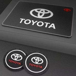 May include: Black rectangular car mat and two round coasters featuring the Toyota logo in white. The rectangular mat has the Toyota logo and text in white. The coasters have the Toyota logo and text in red.