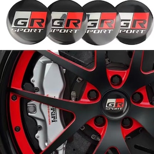 May include: Four black and silver GR Sport wheel centre caps with red accents. The image also shows a close-up of a car wheel with black spokes, red trim, and a silver brake caliper.
