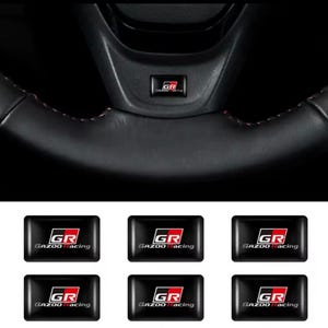 May include: Black steering wheel with red stitching and a small black emblem with the letters "GR" and the words "GAZOO Racing" in white and red. Six additional "GR GAZOO Racing" emblems are below.