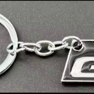 May include: A silver keychain with a rectangular emblem featuring the letters "GR". The emblem is divided into black and red sections, with the letters in white. The keychain includes a silver ring and chain.